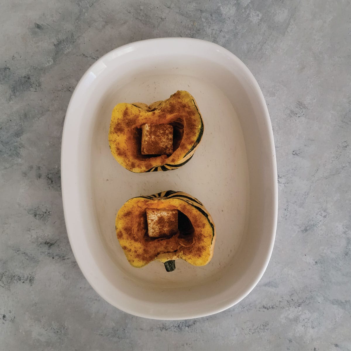 Squash cut in half in a baking dish with a slice of butter in the cavity where the seeds were and spices sprinkled on all over the top.