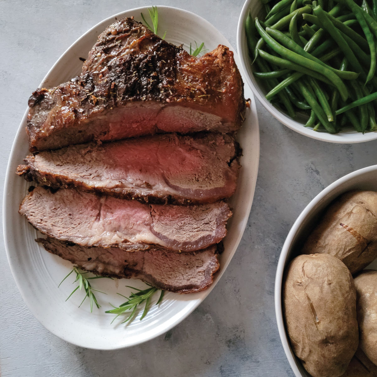 NY strip roast on a platter ready to be served.  Potatoes and green beans in bowls to be served with it.