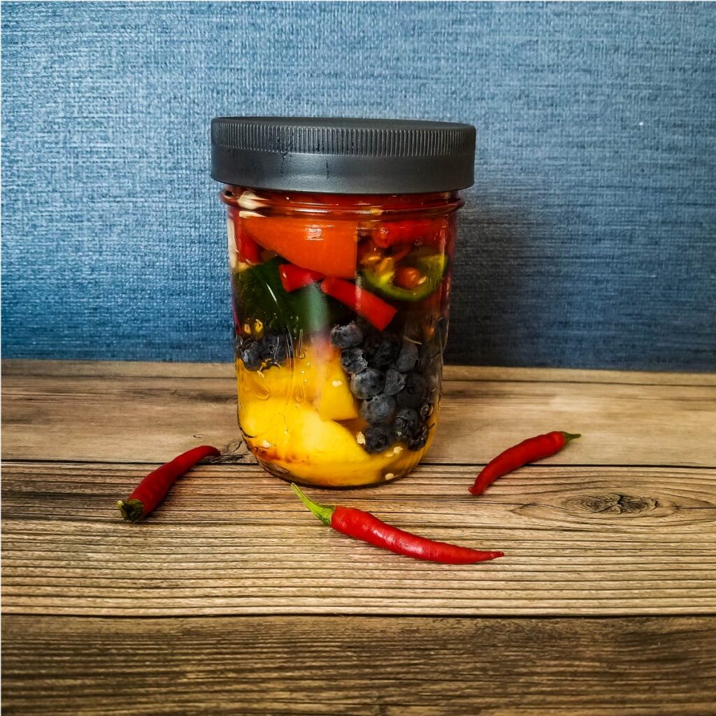 A mix of fruits and vegetables along with hot peppers in a glass jar with a fermenting lid