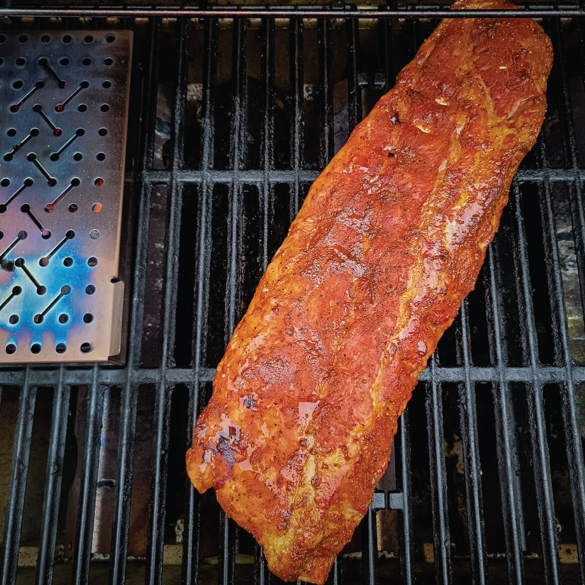 Ribs on a grill cooking with a smoke box next to them.