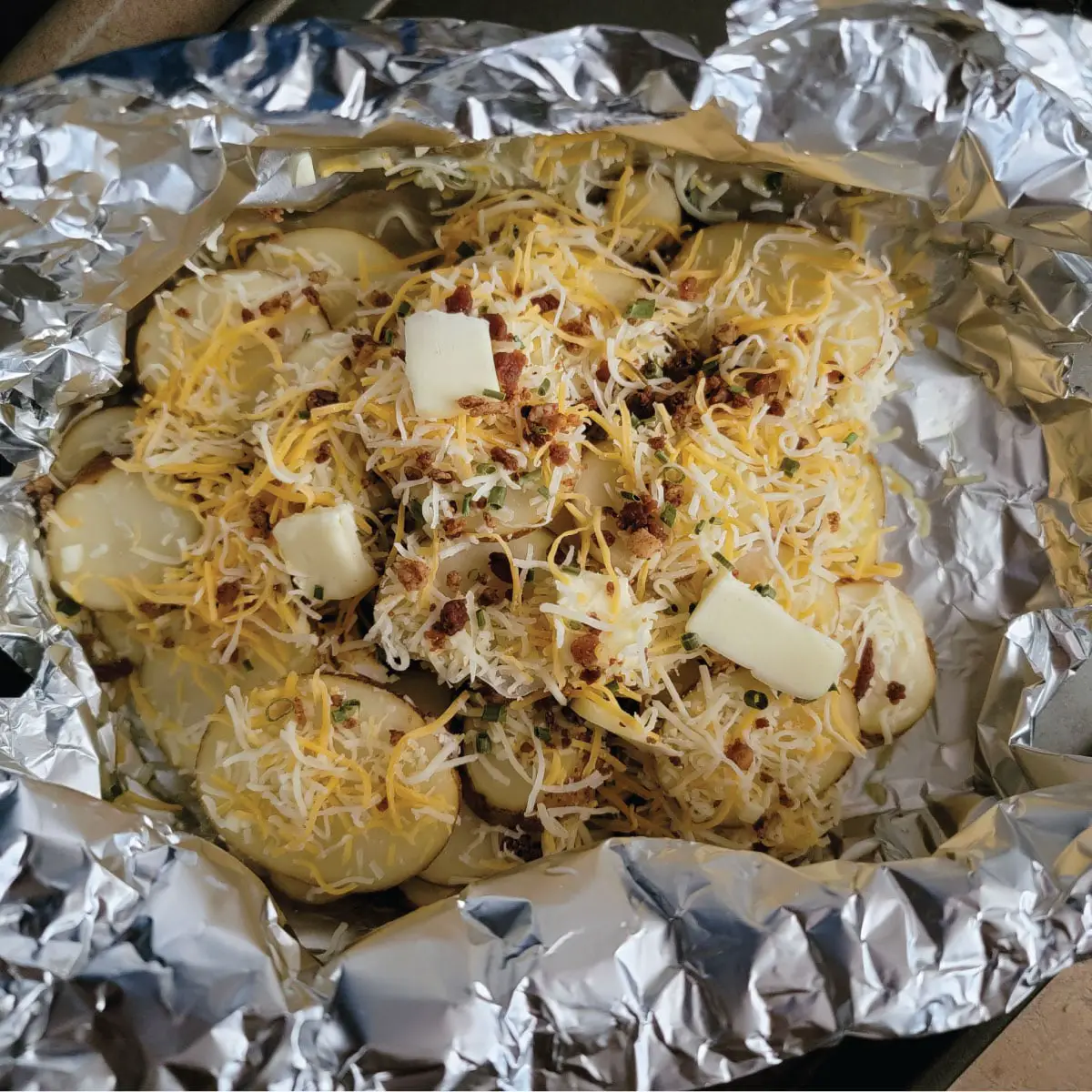 Two layers of potato slices with cheese, bacon, butter and chives on each layer ready to be closed up and put on grill.