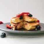 Pancakes on a plate topped with blueberries and strawberries while maple syrup is being poured over them