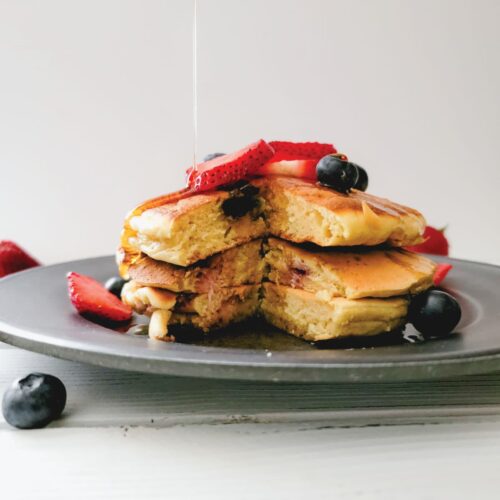 Pancakes on a plate topped with blueberries and strawberries while maple syrup is being poured over them