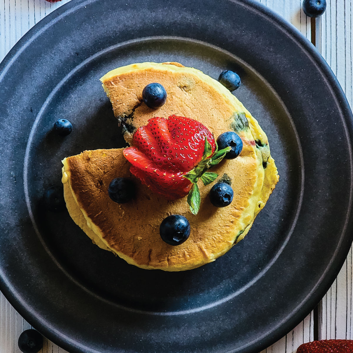 Pancakes on a plate with a wedge cut out of them topped with blueberries and a cut strawberry