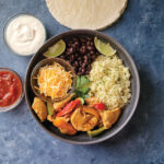 A dinner bowl with chicken fajita filling, rice, beans and cheese. Salsa and sour cream in side dishes.