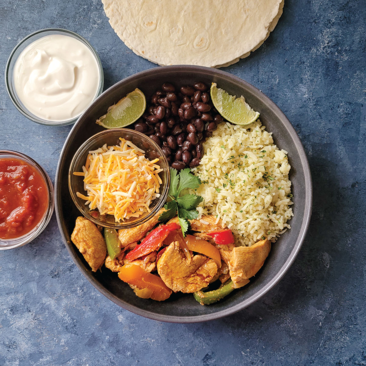 A dinner bowl with chicken fajitas, rice, beans and cheese.  Salsa and sour cream in bowls on the side.