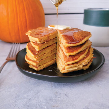 Stack of pumpkin pancakes on a plate with butter on top and maple syrup pouring over the top.