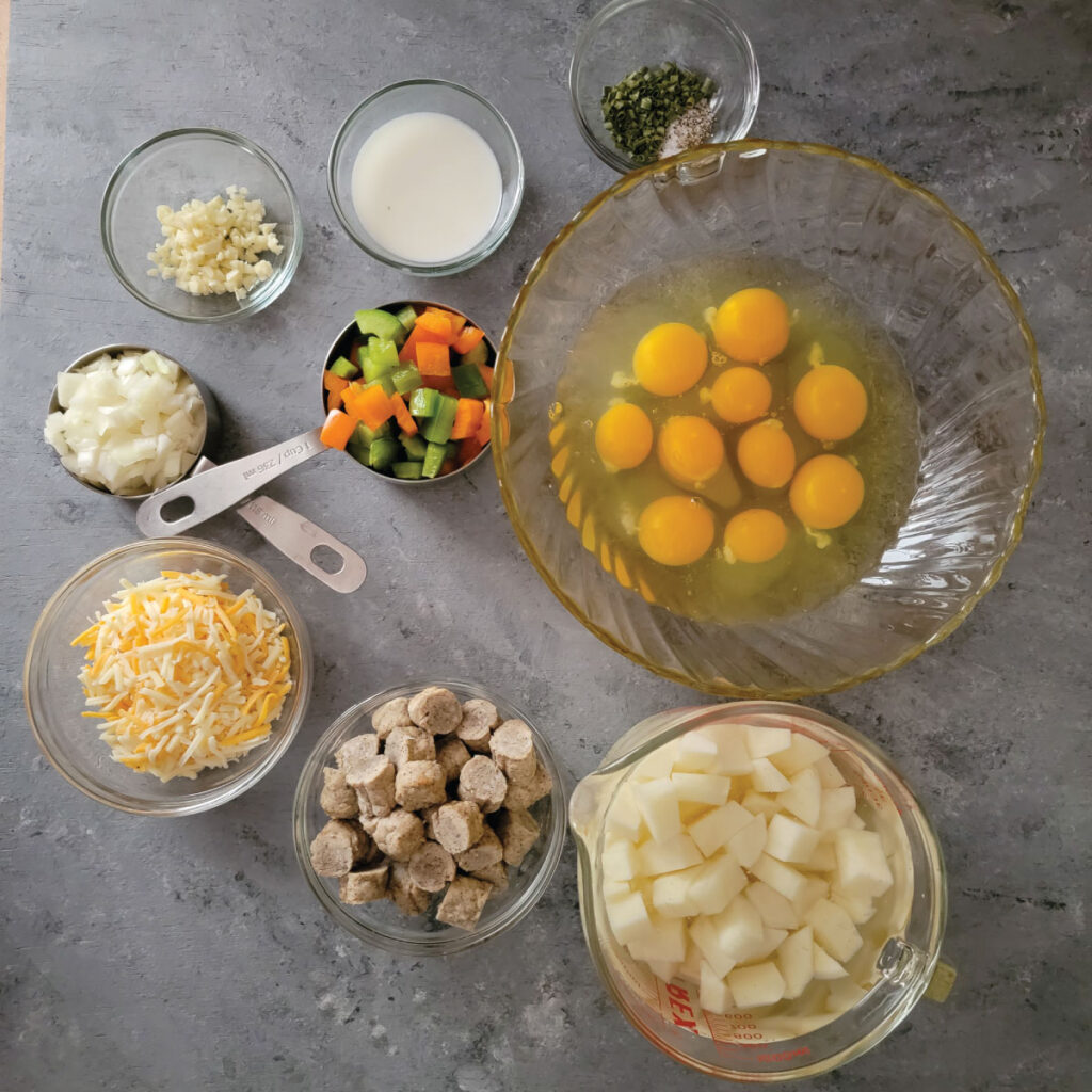 Ingredients for the casserole in prep bowls - eggs, salt, pepper, chives, milk, garlic, onion, peppers, sausage, cheese and potatoes.