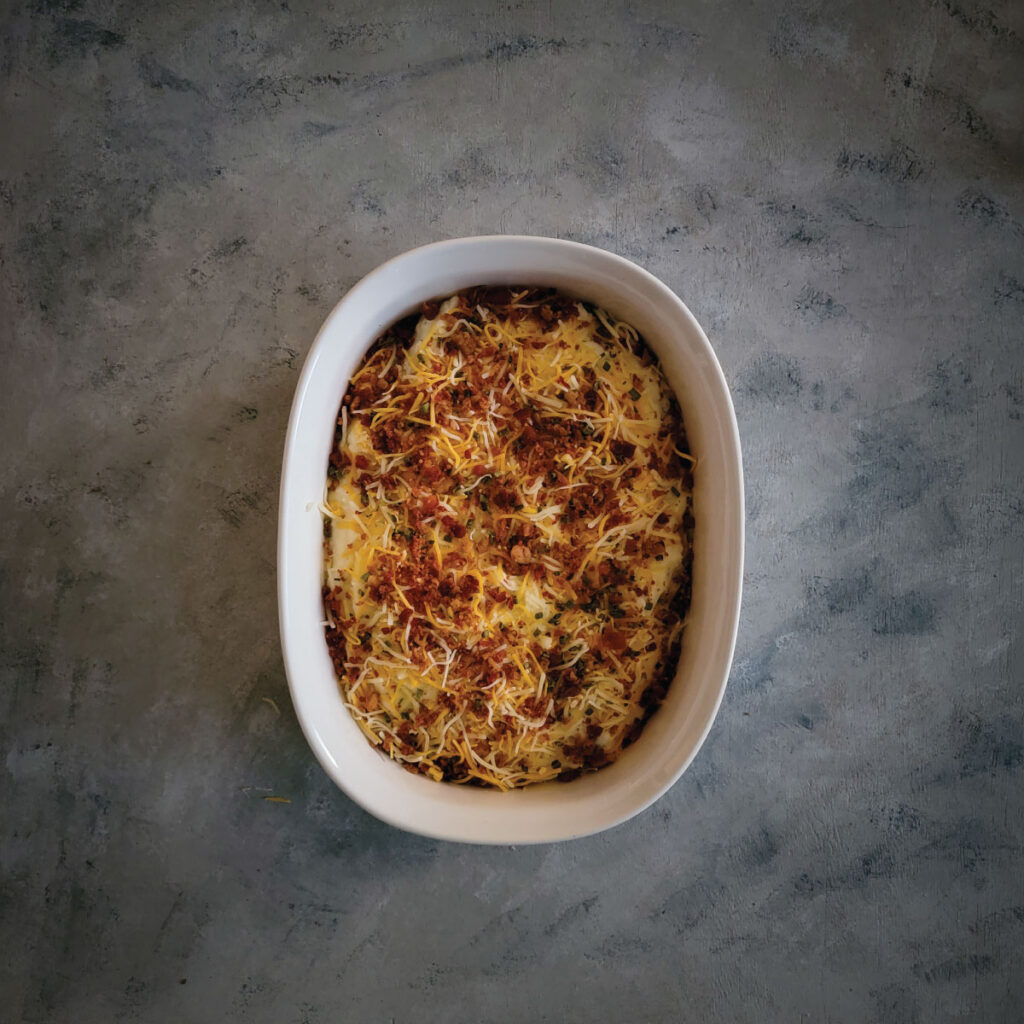 Potatoes mashed with cheeses, sour cream, milk, butter and seasonings in a baking dish with shredded cheese and bacon pieces on top ready to bake.