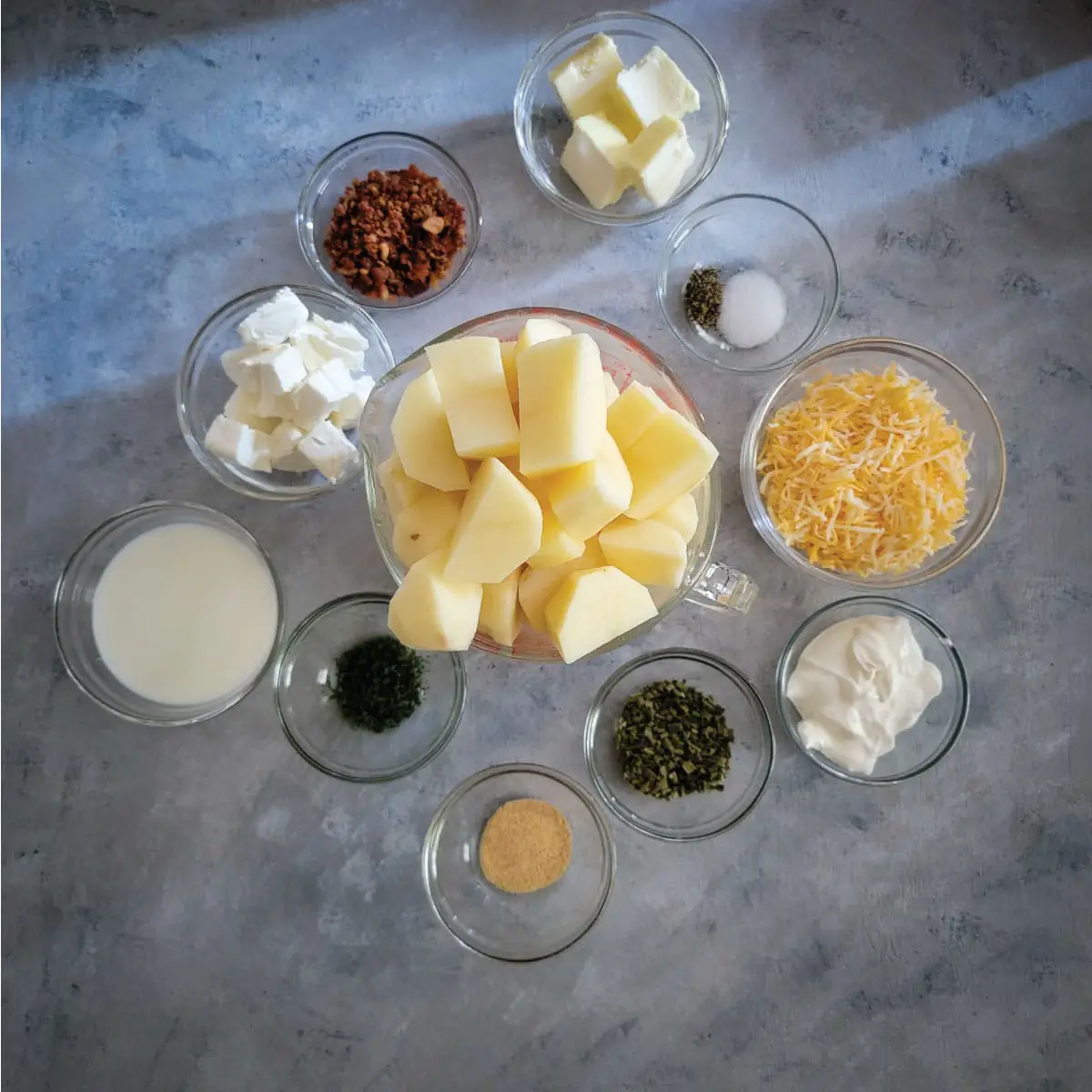 Ingredients in prep bowls - butter, potatoes, salt, pepper, shredded cheese, sour cream, chives, garlic powder, parsley, milk, cream cheese, bacon pieces.