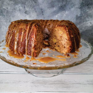 Apple cake with caramel drizzled over the top on a cake platter with a piece of cake cut out to show the apples on the inside.