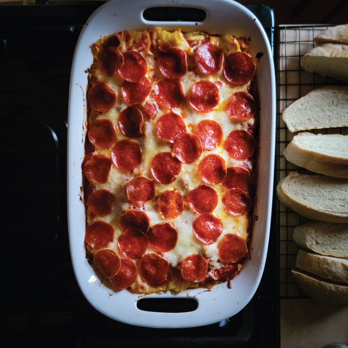 Lasagna out of the oven resting on the stove before serving with sliced Italian bread.