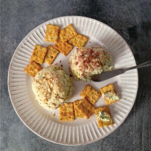 2 cheeseballs on a plate with crackers spread around it. 2 of the crackers have some of the cheese on them ready to eat.