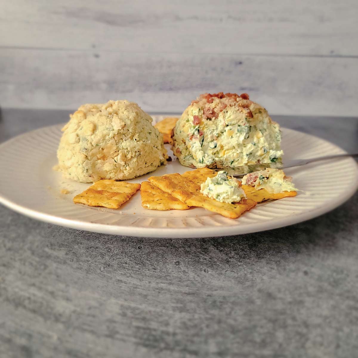 2 cheeseballs on a plate with crackers spread around it. 2 of the crackers have some of the cheese on them ready to eat.