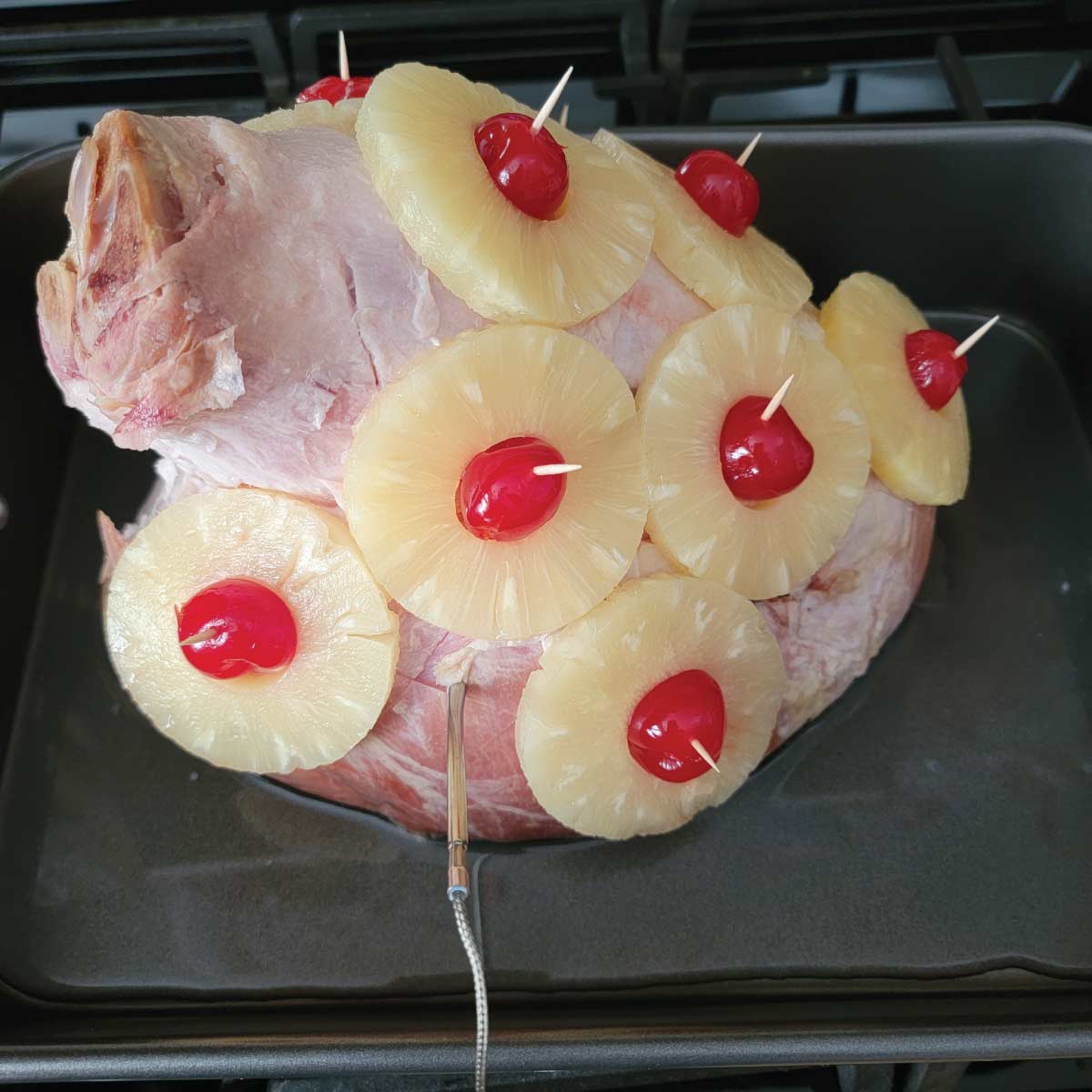 Ham in a roasting pan with pineapple slices and cherries on it with a thermometer probe in it.