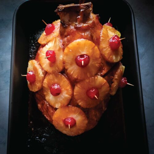 Baked ham in a roasting pan with pineapple slices and cherries on it resting after cooking and before cutting.