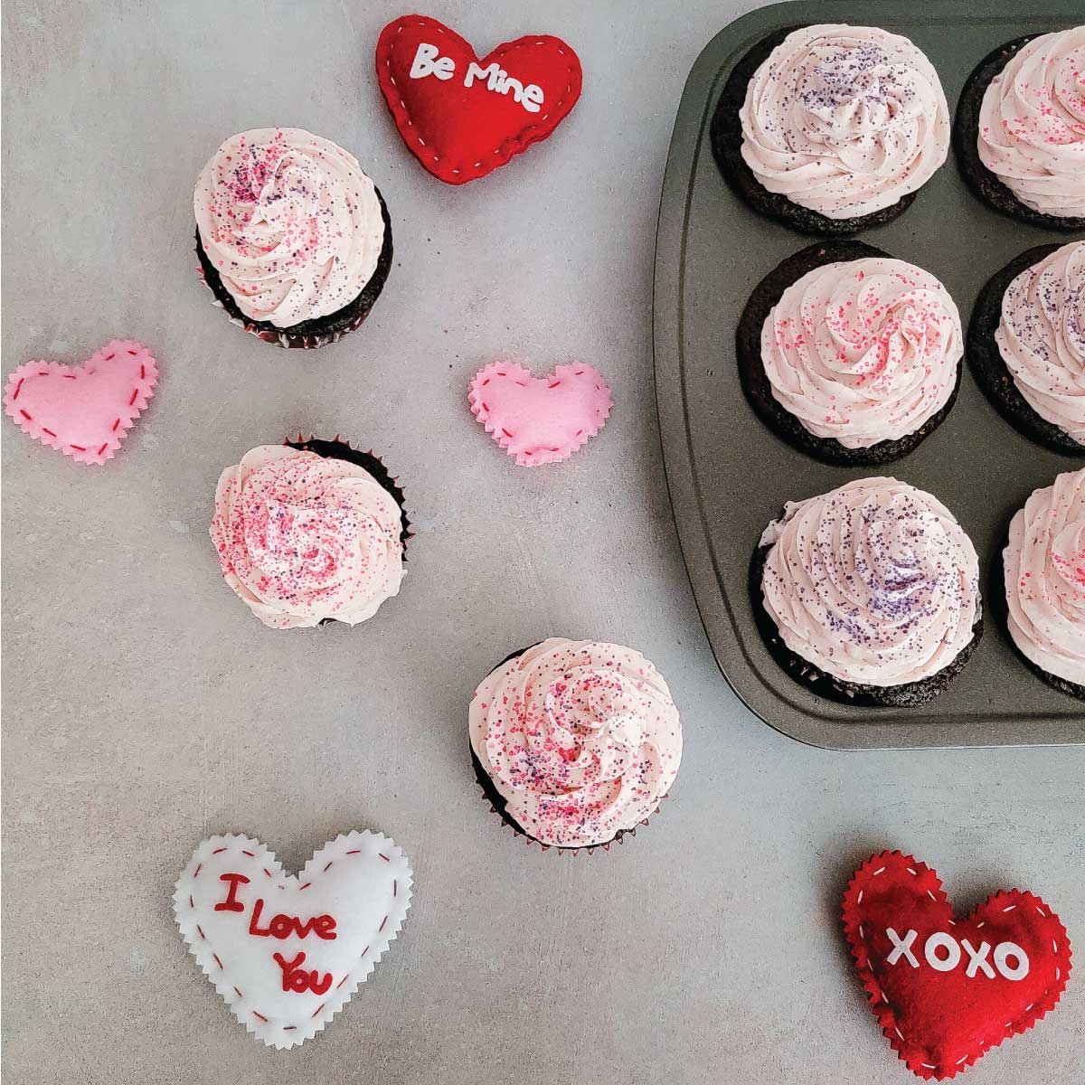 Cupcakes with icing swirled on top and decorative sugar sprinkled on top with felt hearts scattered around for Valentine's Day decorations.
