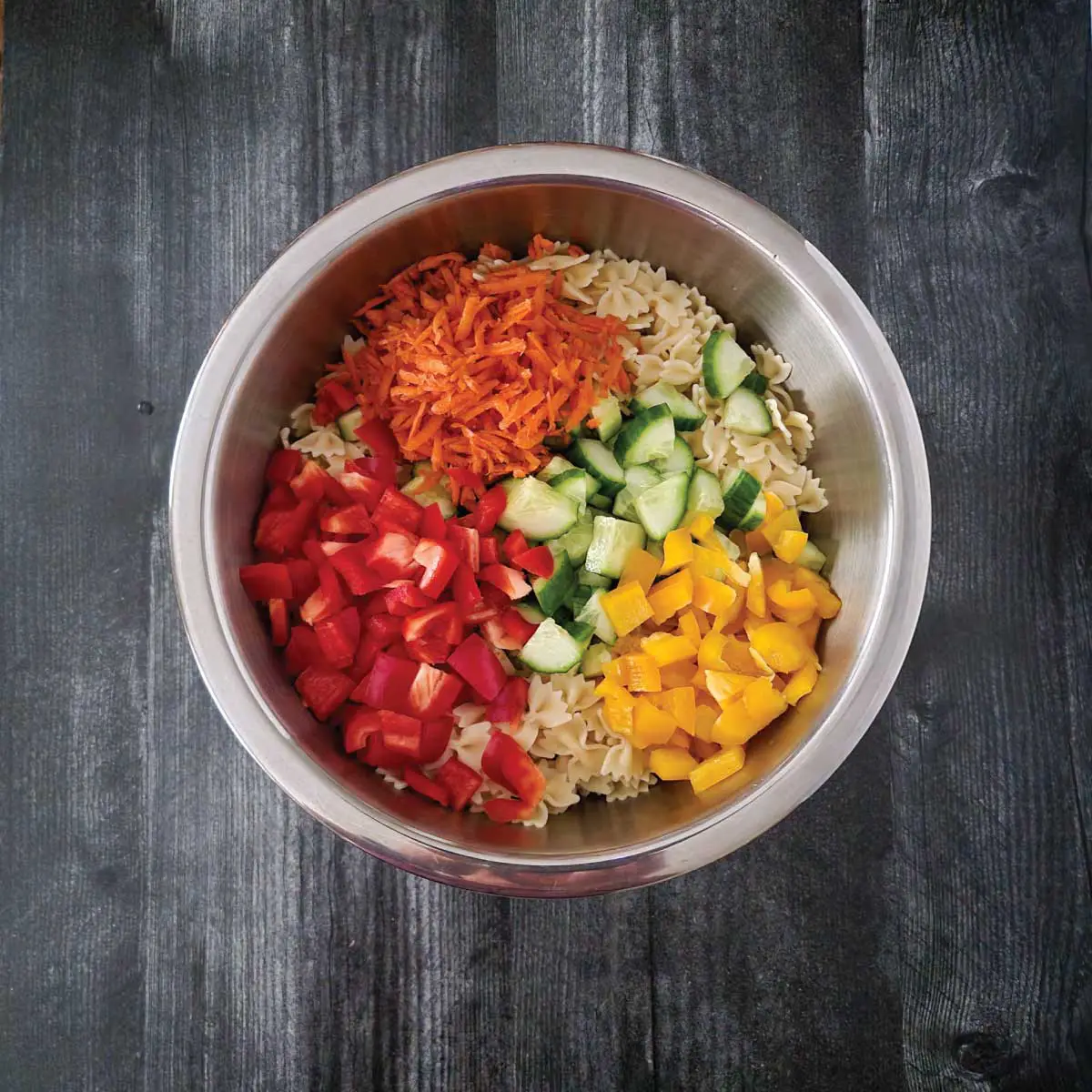 Noodles in a large mixing bowl with chopped red and yellow peppers, cucumbers and shredded carrot on top ready to be mixed.