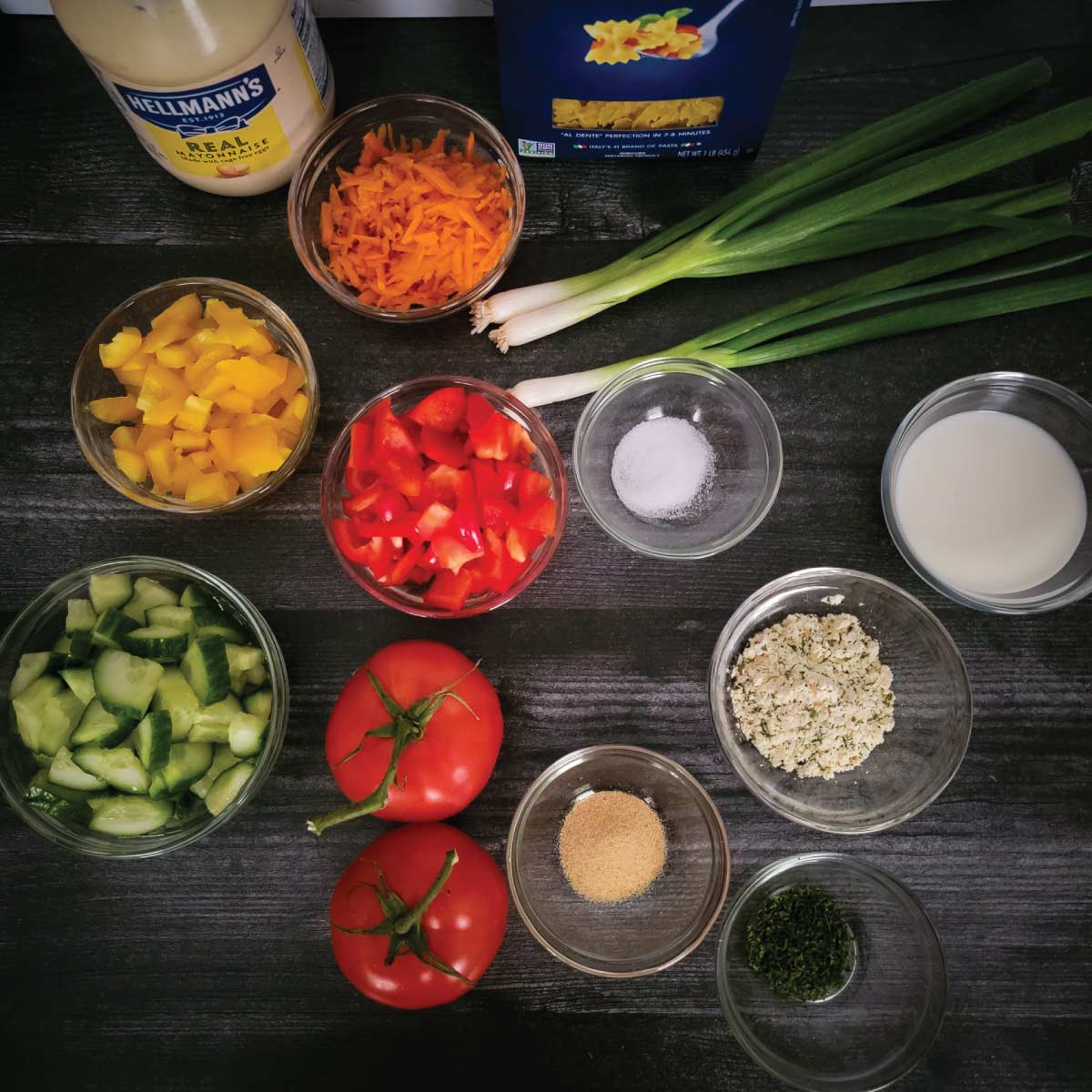 Ingredients prepped for the pasta salad - mayonnaise, pasta, shredded carrot, scallions, milk, salt, chopped red pepper, chopped yellow pepper, cut cucumber, tomatoes, garlic powder, ranch seasoning parsley for garnish.
