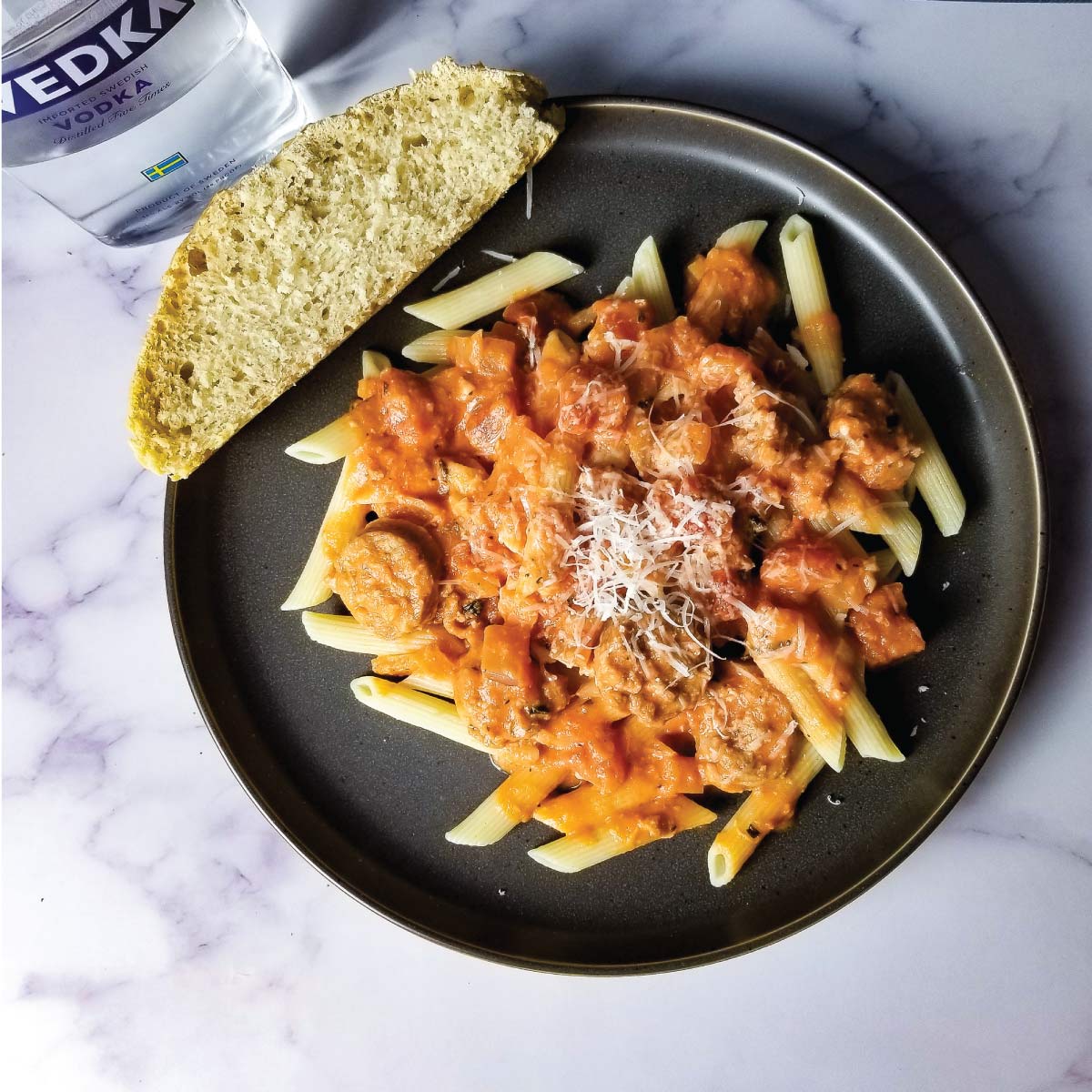 Penne pasta on a plate topped with vodka sauce and a slice of Italian bread on the side.