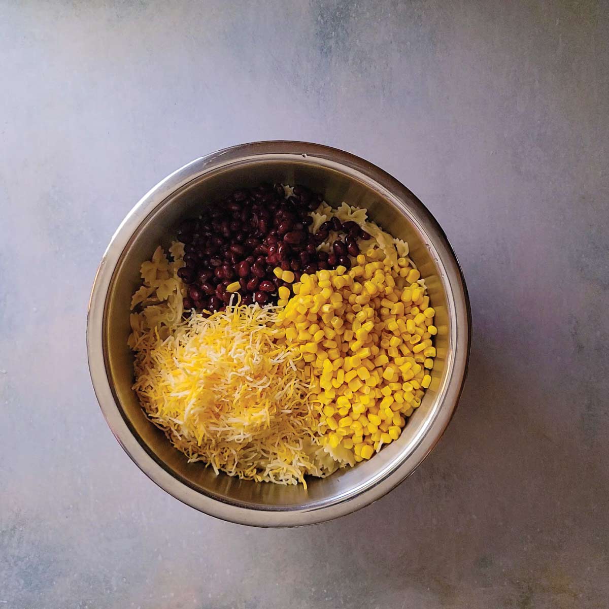 Noodles, black beans, corn and shredded cheese in a mixing bowl ready to stir together.