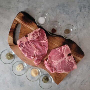 2 t-bone steaks on a cutting board with the seasonings prepped in small bowls to display the ingredients.