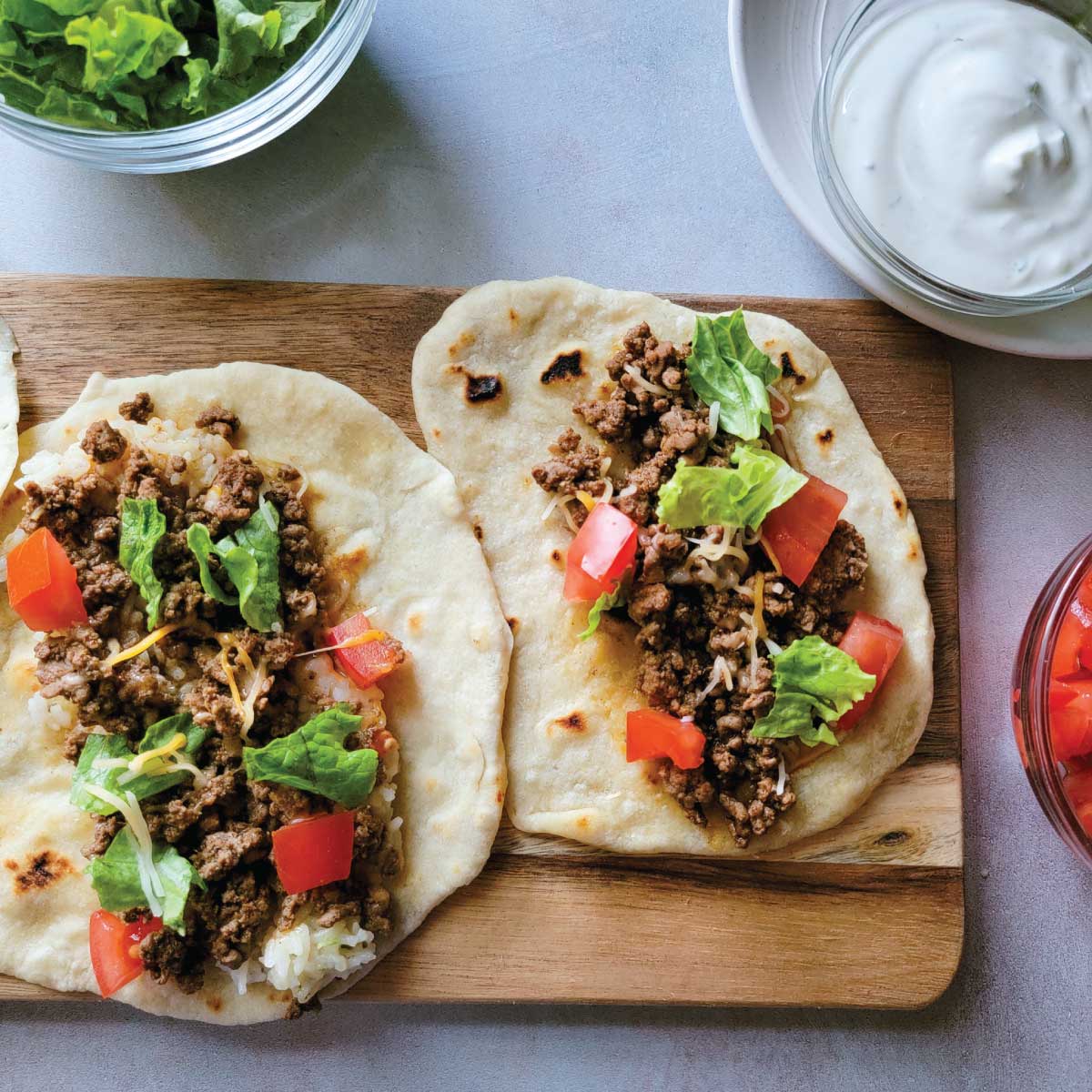 Soft tacos made with homemade tortillas on a serving board with diced tomatoes, cut lettuce, lime wedges, sour cream and taco sauce in bowls for toppings.