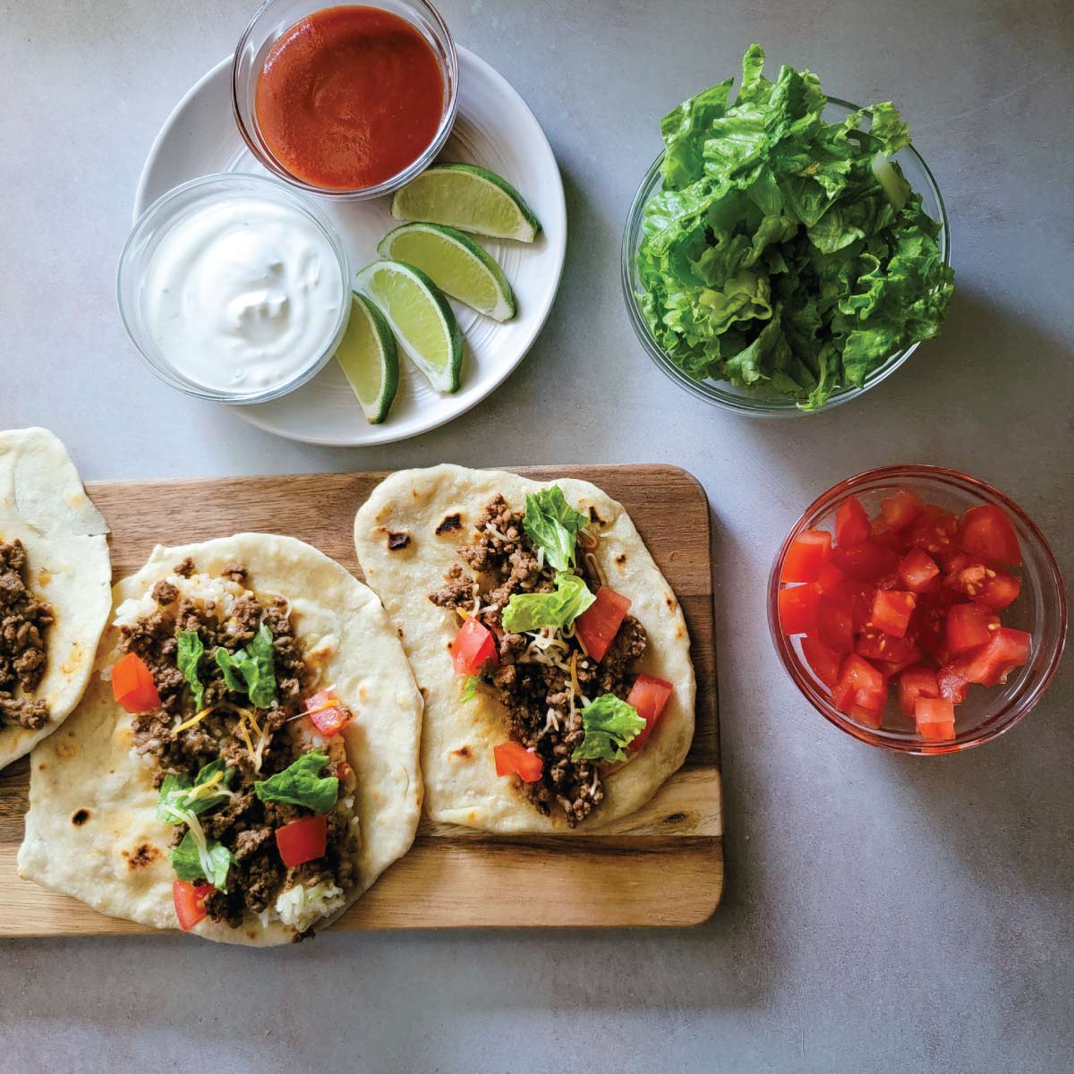 Soft tacos made with homemade tortillas on a serving board with diced tomatoes, cut lettuce, lime wedges, sour cream and taco sauce in bowls for toppings.