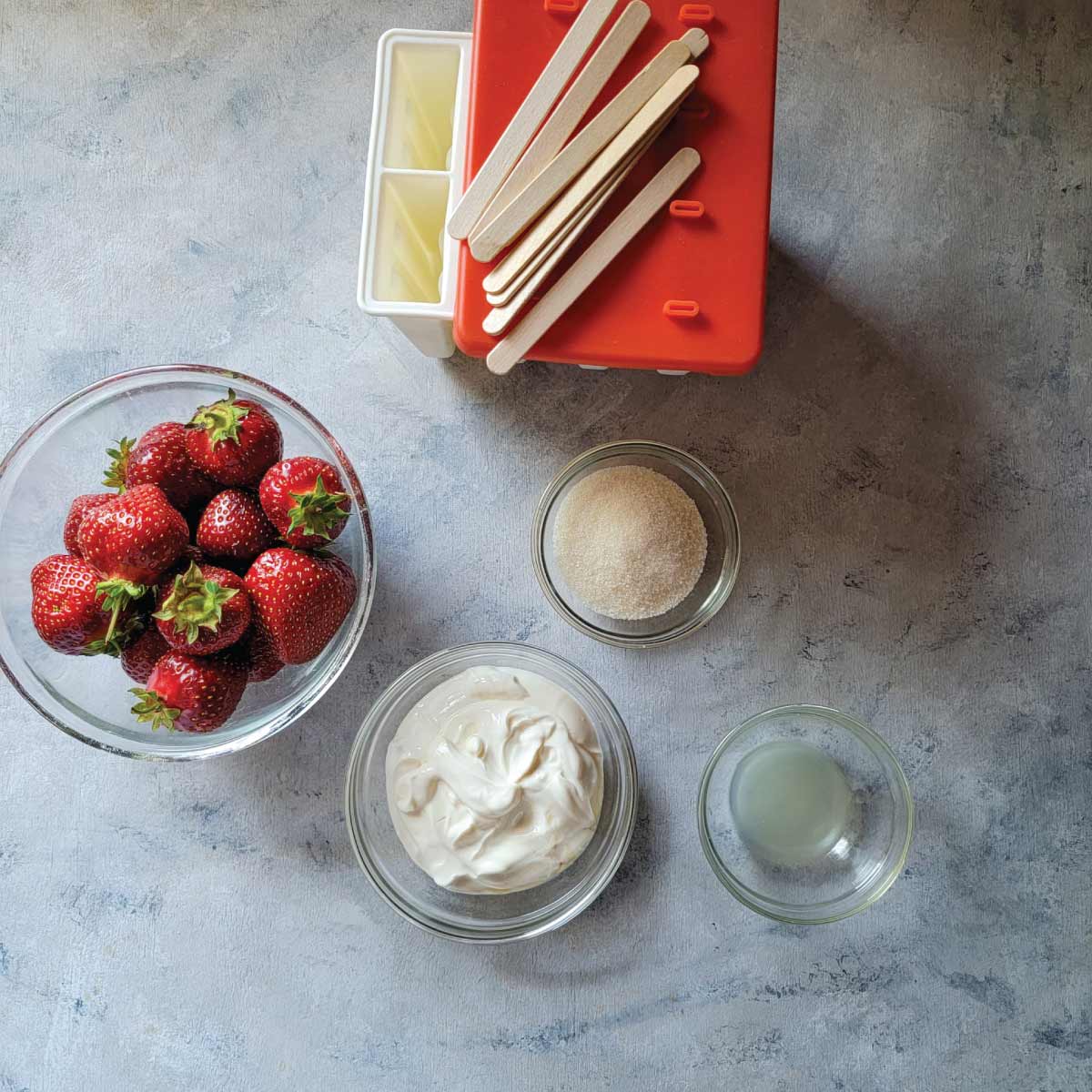 Strawberries, yogurt, sugar and lemon juice in prep bowls with a silicone popsicle mold ready to make popsicles.