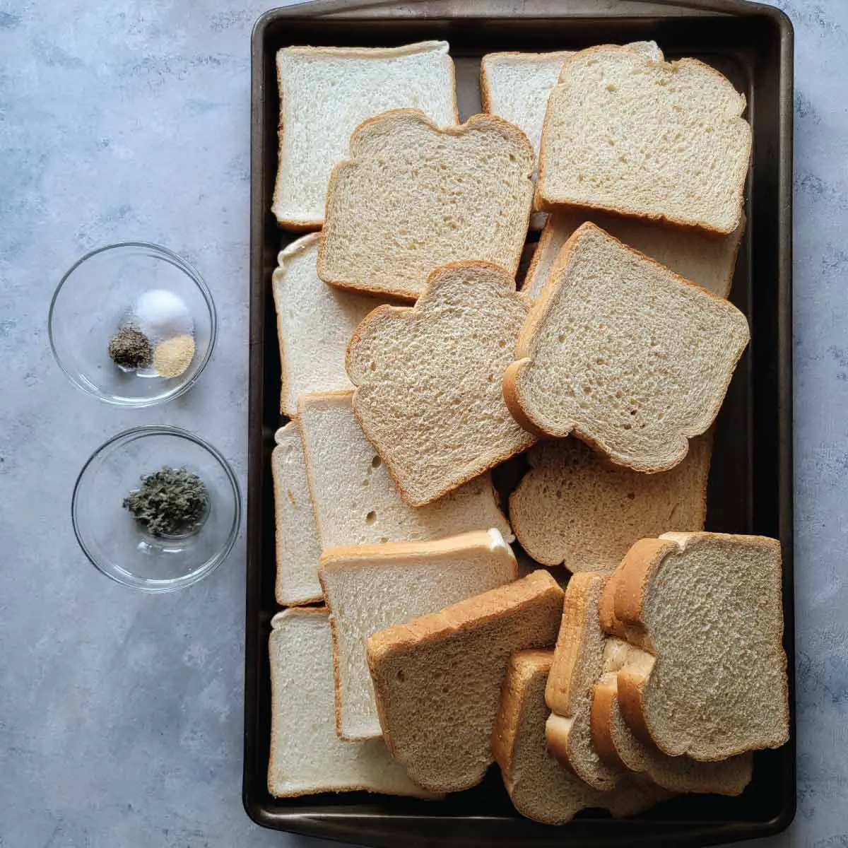 White and wheat bread slices on a baking tray along with the seasonings in prep bowls.