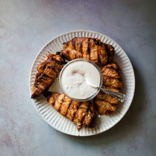 4 grilled chicken breasts on a plate with a bowl of creamy garlic sauce in the middle of the plate for serving.