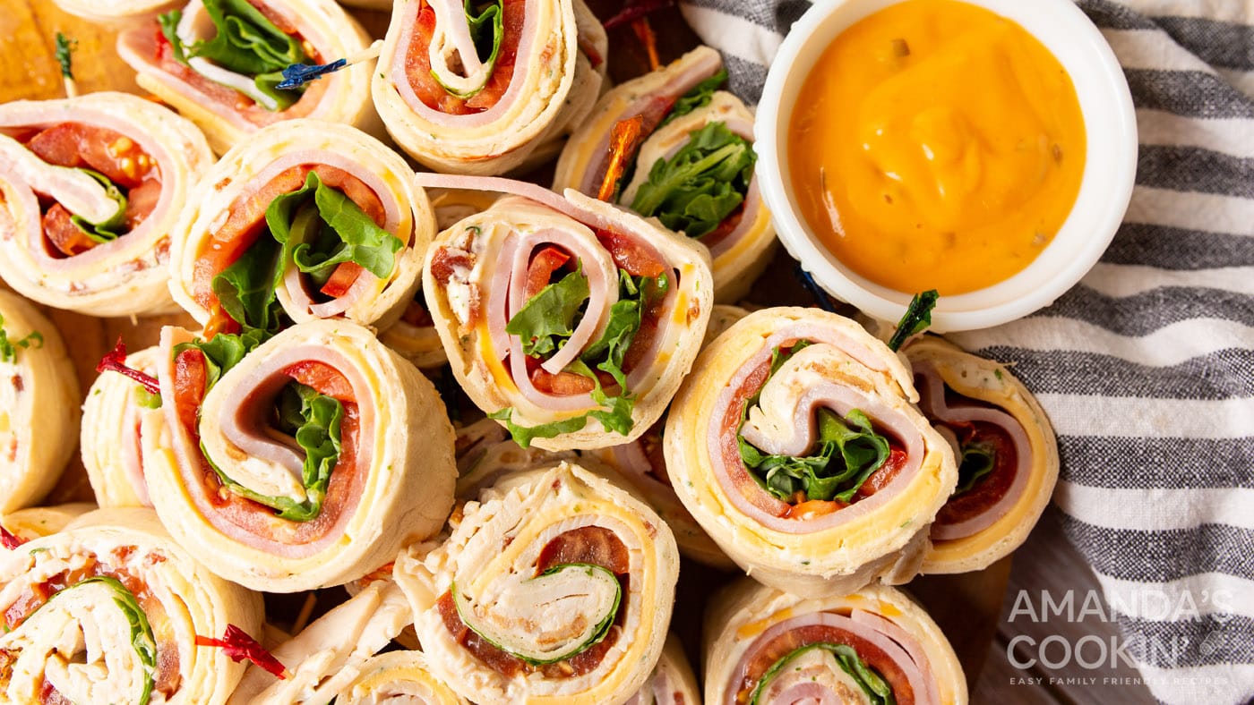 Pinwheel sandwiches stacked on a plate with a dipping sauce in a small prep bowl on the side.