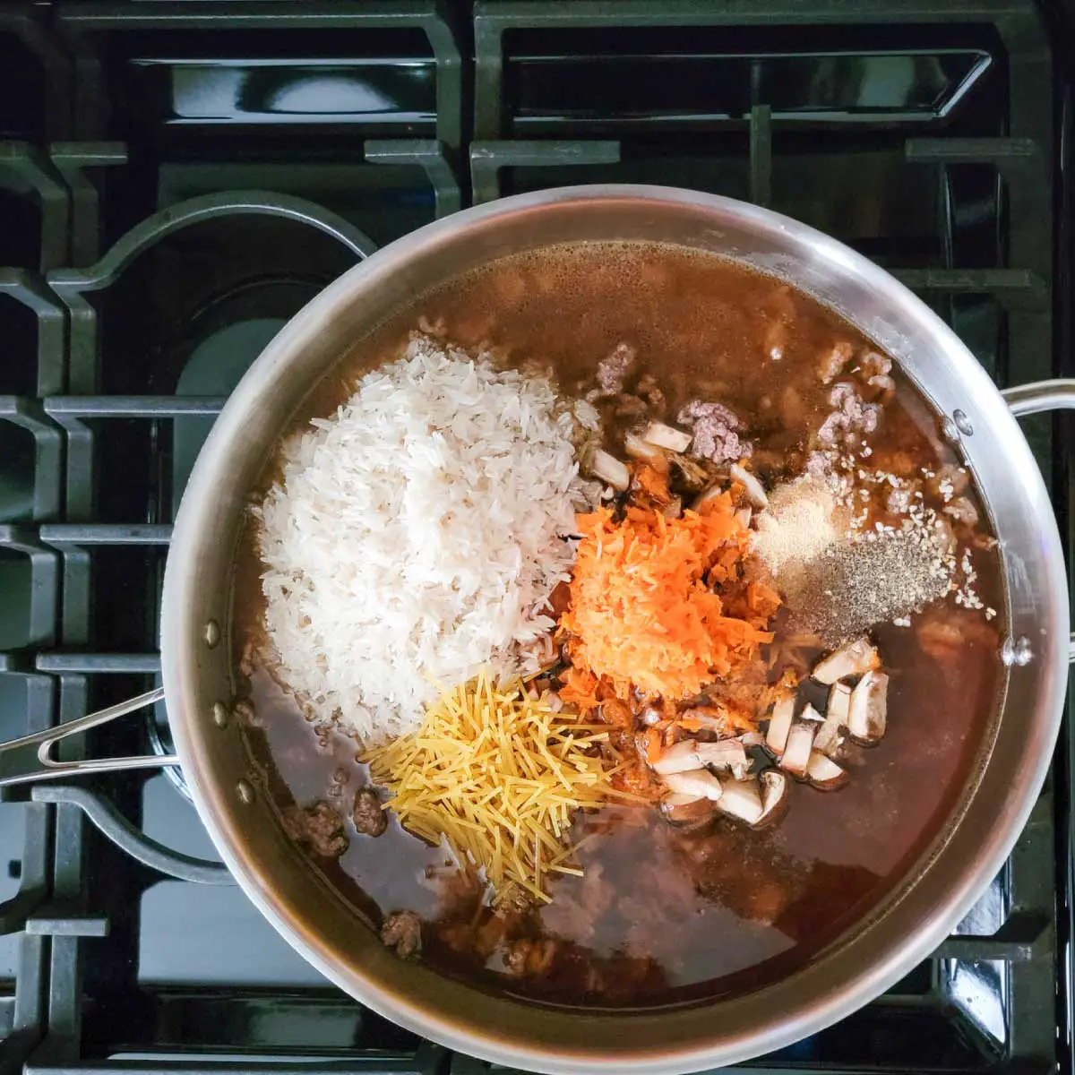 Ground beef, rice, pasta, beef broth, carrot, mushrooms and spices in a large frying pan ready to be stirred and cooked.