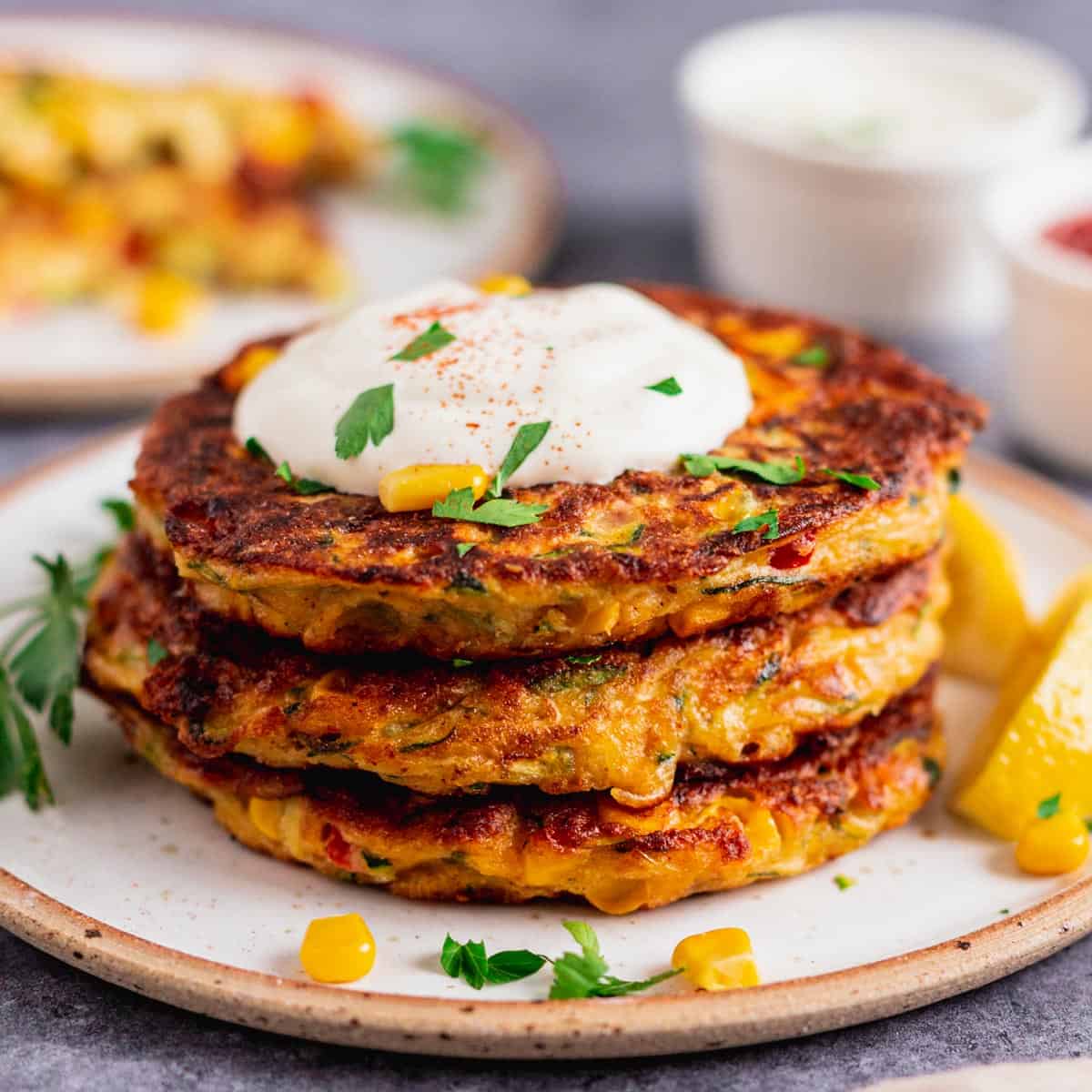 3 zucchini fritters stacked on a plate topped with sour cream and fresh parsley as a garnish.