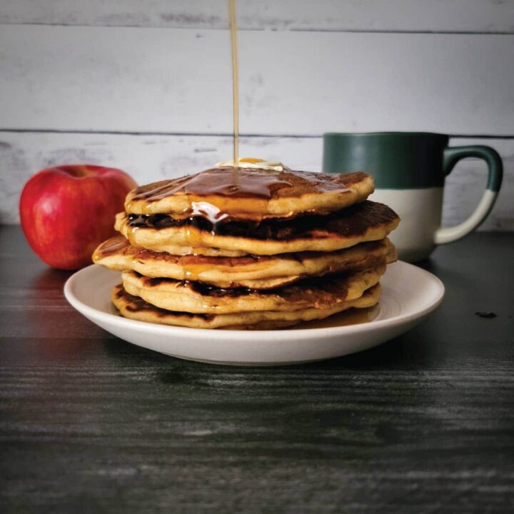 Apple pancakes stacked on a plate with maple syrup being drizzled over the top.