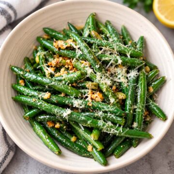 Garlic butter green beans in a white serving bowl with perfectly browned garlic and fresh shredded parmesan as a topping.